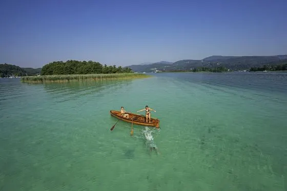 Zwei Personen in einem Holzboot auf dem türkisgrünen Wörthersee, dahinter eine bewaldete Insel und die Berge